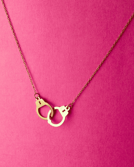 Colgante de esposas dorado en acero inoxidable sobre fondo rosa. Joya con actitud urbana y acabado en oro brillante.