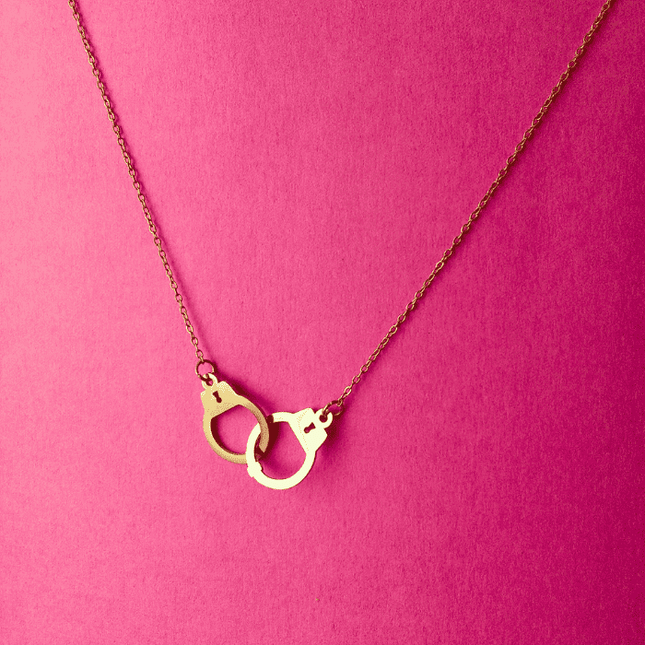 Colgante de esposas dorado en acero inoxidable sobre fondo rosa. Joya con actitud urbana y acabado en oro brillante.
