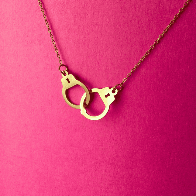 Detalle del colgante en forma de esposas entrelazadas doradas. Diseño simbólico con un toque de rebeldía y glamour.