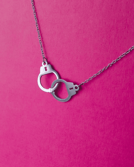 Colgante de esposas plateado en acero inoxidable sobre fondo rosa. Diseño provocador y urbano con acabado brillante.