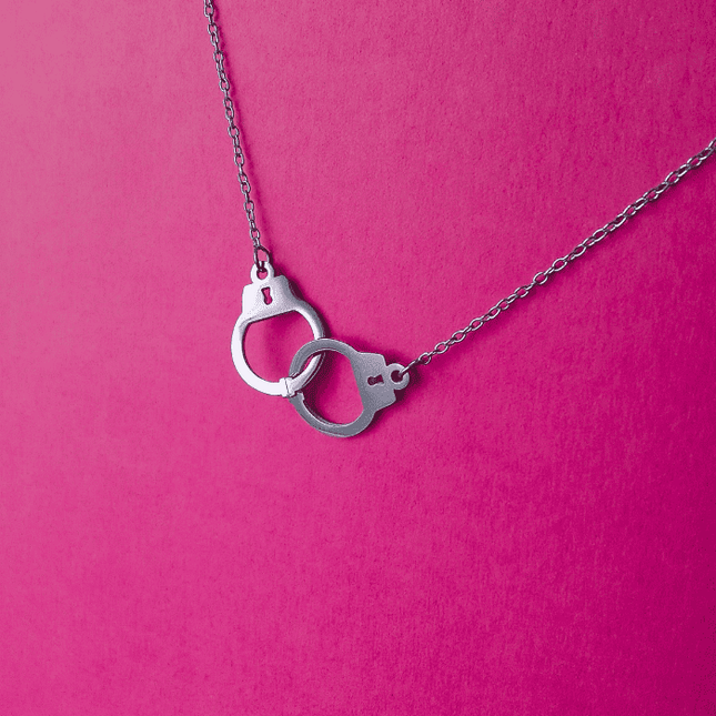 Colgante de esposas plateado en acero inoxidable sobre fondo rosa. Diseño provocador y urbano con acabado brillante.