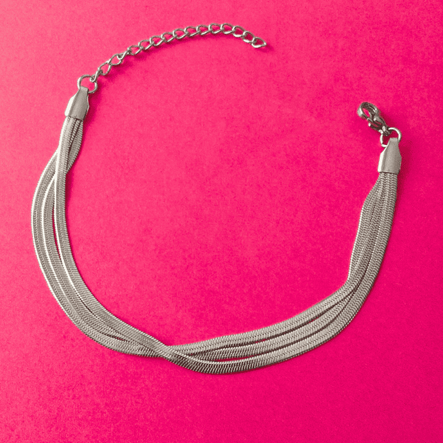 Pulsera plateada de triple tira trenzada en acero inoxidable sobre fondo rosa. Diseño elegante y moderno que refleja la luz.